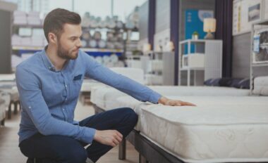 Young handsome man examining orthopedic mattress on sale at furniture store, copy space. Attractive male customer buying new bed at homeware supermarket. Consumerism, home concept